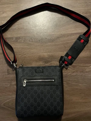 Svart axelväska - Snygg svart axelväska från med GG-monogram över hela väskan. Väskan har en justerbar axelrem i svart och rött, en framficka med dragkedja och detaljer i silver. Perfekt för dig som vill ha en stilren och lyxig accessoar.