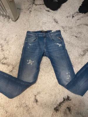 Blå slitna jeans från Replay - Säljer ett par blå jeans från Replay med slitningar och distressed detaljer på både fram- och baksida. Klassisk femficksmodell med smal passform och läderpatch med Replay-logga bak i midjan. Perfekta för en avslappnad och trendig look.