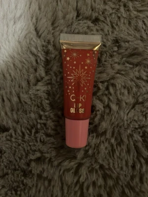 GKI Lip Gloss med stjärndesign - Säljer ett läppglans från GKI i en snygg tub med rosa och guldiga detaljer samt stjärnmönster. Färgen är en varm rosa nyans och förpackningen har en praktisk applikator. Perfekt för dig som vill ha glansiga och återfuktade läppar.