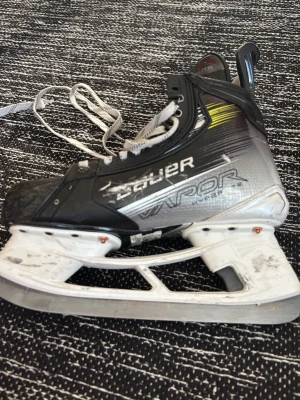 Bauer Vapor hockeyskridsko - Säljer en Bauer Vapor hockeyskridsko i svart, silver och vitt med gula detaljer. Skridskon har snörning, robust plös och klassisk form. Bladet är i stål och skon är tillverkad i syntetmaterial för extra hållbarhet. Användes bara i en säsong 