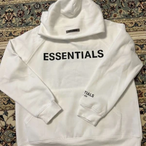  Essentials hoodie - Säljer en clean vit hoodie från Essentials Fear of God med svart tryck på bröstet och ärmen. Klassisk oversized passform, stor huva och mjukt material. Perfekt för dig som gillar streetwear och vill ha en stilren look. Storlek M men passar även L. Är lite står för mig i och med att jag använder storlek S.