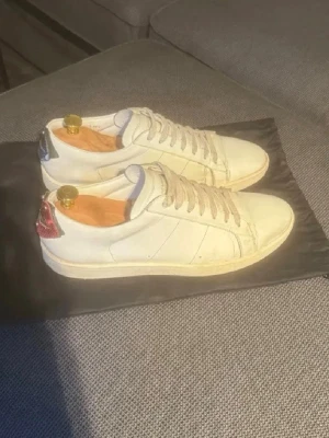 Saint Laurent vita sneakers med detaljer - Säljer ett par vita Saint Laurent sneakers i läder med klassisk låg profil. Skorna har kontrasterande hälflikar, en i rött och en i blått, samt vita snören och diskreta sömdetaljer. Perfekta för dig som gillar stilrena och exklusiva sneakers. Strl 43 men passar defenetivt 44