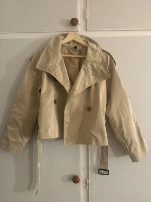 Virala trenchcoat från H&M  - Kort trenchcoat från H&M  med bred krage, dubbelknäppning och bälte i midjan. Ena ärmremmen saknas, men jackan går fortfarande utmärkt att använda precis som den är. Det går säkert även att köpa liknande remmar i butik om man vill. Fråga gärna om bilder