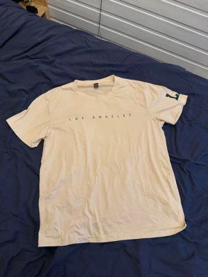Beige t-shirt med LA-tryck - Snygg beige t-shirt med texten 'LOS ANGELES' tryckt över bröstet och ett stort grönt 'LA' på ena ärmen. Klassisk rund hals och korta ärmar. Perfekt för en avslappnad streetwear-look.