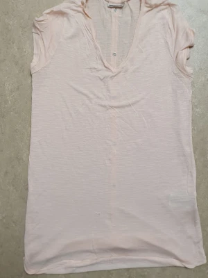 T-shirt - Säljer en ljusrosa t-shirt med v-ringning och korta ärmar. T-shirten har knappar längs hela ryggen och är gjord i ett tunt, mjukt bomullstyg. Perfekt för dig som gillar en enkel men ändå snygg stil.