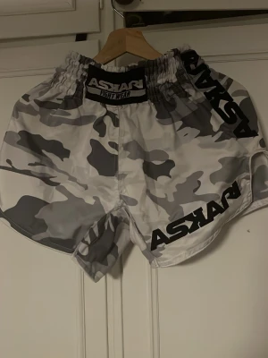 Camo shorts från Askari Fight Wear - Snygga träningsshorts från Askari Fight Wear i grått och vitt camouflagemönster. De har elastisk midja och är tillverkade i ett lätt och slitstarkt syntetmaterial, perfekta för intensiva träningspass.