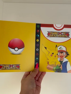 Pokémon Next Destinies samlarpärm - Säljer en färgglad Pokémon Next Destinies samlarpärm med gul framsida, motiv av Ash och Pikachu samt Pokéboll. Pärmen har plastfickor för att förvara och organisera samlarkort. Perfekt för Pokémonkort och andra samlarkort. Plats för 240 st kort 