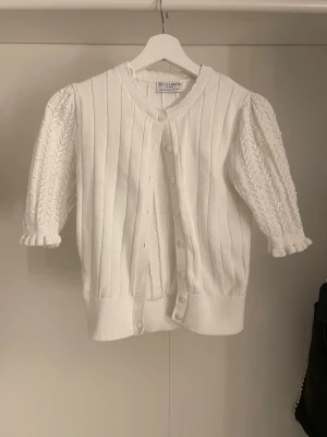 Vit kortärmad kofta - Söt vit kofta från Holly & White med puffärmar och fina virkade detaljer på ärmarna. Koftan har knappar framtill, ribbade muddar och en mjuk, stickad struktur. Köpt för 500kr.