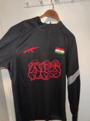 Svart långärmad topp med tryck - Svart långärmad topp från Joma med halv dragkedja framtill. Framsidan har ett rött tryck med tre apor, ett gevär och en flagga. Reflekterande detaljer på ärmarna. Perfekt för dig som gillar unika plagg med statement-tryck.