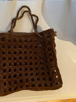 Brun virkad väska - Unik brun virkad nätväska med rutigt, öppet mönster. Väskan har dubbla handtag och är gjord i mjukt garn, vilket gör den flexibel och lätt att bära. Perfekt för dig som vill ha en trendig accessoar med handgjord känsla.