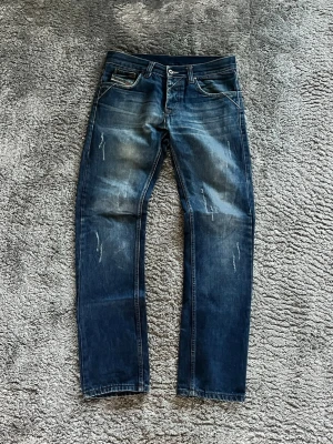 Blå slitna jeans från Diesel W30/L32 - Säljer ett par blå jeans från Diesel med snygga slitningar och tvättade detaljer. Klassisk femficksmodell med raka ben och låg midja. Jeansen har en cool, avslappnad look och är tillverkade i bomull med lite stretch för extra komfort. 