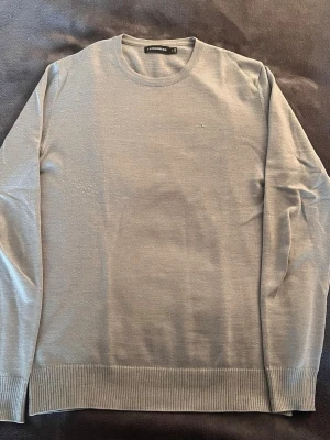 Ljusgrå tröja från J.Lindeberg - Ljusgrå, tunnstickad tröja från J.Lindeberg med rund hals och diskret broderad logga på bröstet. Tröjan har ribbade muddar vid ärmslut och nederkant och är tillverkad i ett mjukt material som ger en clean och stilren look.