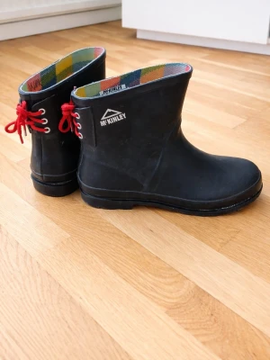 Svarta McKinley Tindra gummistövlar - Svarta gummistövlar från McKinley, modell Tindra, med färgglatt rutigt foder och röda snören bak. Perfekta för regniga dagar och har en klassisk, stilren design. Tillverkade i slitstarkt gummi och fodrade för extra komfort.