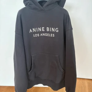 Svart hoodie från Anine Bing - Snygg svart hoodie från Anine Bing med vit text 'ANINE BING LOS ANGELES' på bröstet. Klassisk passform med huva och känguruficka framtill. Tillverkad i 100% bomull för en mjuk och bekväm känsla. Perfekt för en avslappnad och trendig look.