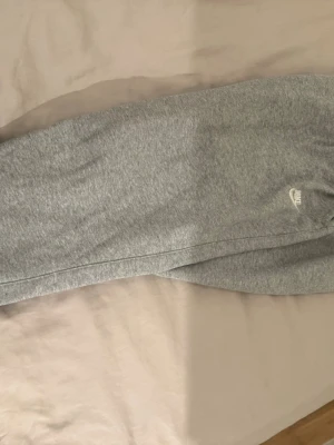 Gråa mjukisbyxor från Nike - Säljer ett par klassiska gråa mjukisbyxor från Nike med dragsko i midjan och diskret Nike-logga på låret. Byxorna har en avslappnad passform och är tillverkade i mjukt sweatshirtmaterial som är perfekt för chill dagar.