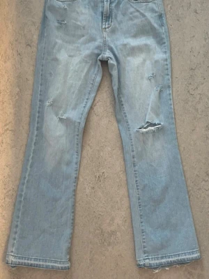 Ljusblå bootcut jeans med slitningar - Säljer ett par ljusblå bootcut jeans med trendiga slitningar. Jag har själv förlängt benen längst ned, genom att sprätta upp fållen. Jag har dessutom skapat slitage och hål (se bilder) för att få fram en sliten look. Innerben längd: 72cm.  Storlek 27 (tror storleken gäller midjan). Mid-waist.  Enligt lappen i jeansen är detta ”Johanna kick flare style” med en ”bright lime wash”, de är iallafall i en väldigt fin ljusblå färg. Kan tyvärr inte längre ha dem pga att de har blivit försmå. 