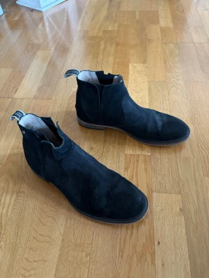 Svarta Chelsea boots i mocka från Sneaky steve - Säljer ett par svarta Chelsea boots från Sneaky steve i mjuk mocka. Klassisk siluett med resår på sidorna och dragflikar med logga bak. Snygg kontrast med brun sula och låg klack. Perfekta för en stilren och avslappnad look.