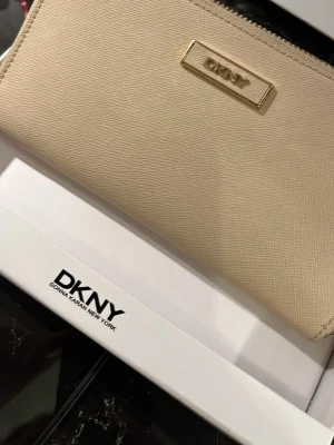 Beige plånbok från DKNY - Snygg beige plånbok från DKNY i stilren design med guldfärgad logotyp framtill. Plånboken är tillverkad i ett strukturerat konstläder och har dragkedja med metall-detalj. Perfekt för att hålla koll på kort och cash.