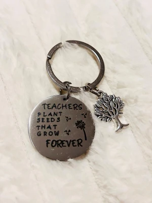 Nyckelring med text och trädberlock - Snygg nyckelring i silverfärgad metall med rund bricka där det står 'Teachers plant seeds that grow forever' och en liten berlock i form av ett träd. Perfekt accessoar för att piffa upp nyckelknippan eller väskan.