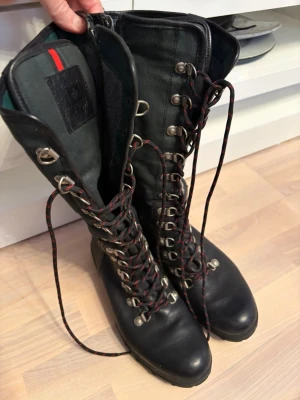 Svarta höga läderkängor Canada Snow - Säljer ett par svarta höga snörkängor från Canada Snow. Kängorna är i läder med robust sula och har röda och svarta snören. Insidan är fodrad för extra värme och komfort. Perfekta för kalla dagar och outdoor-äventyr.