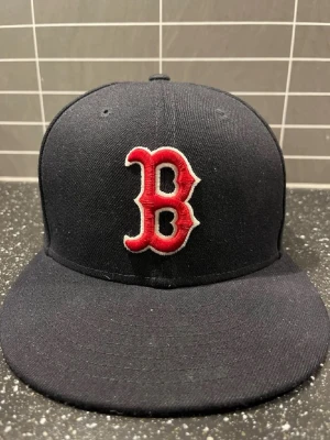 Boston Red Sox svart keps New Era - Snygg svart keps från New Era med Boston Red Sox logga broderad i rött och vitt framtill. Klassisk rak skärm och MLB-logga baktill. Tillverkad i slitstarkt tyg, perfekt för dig som gillar sportig streetstyle.