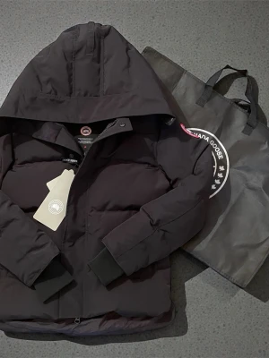 Svart dunjacka från Canada Goose - Svart dunjacka från Canada Goose med huva och klassisk logotyp på ärmen. Jackan har en stilren, quiltad design och ribbade muddar vid ärmslut. Perfekt för kalla dagar och har en snygg, enkel look som funkar till det mesta.