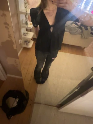 Svart blus med volang och cut out - Säljer en svart blus med korta ärmar och snygg cut out-detalj framtill. Blusen har volang nedtill och v-ringad hals. Perfekt för dig som gillar en trendig och lite edgy stil. Materialet känns lätt och flowy.