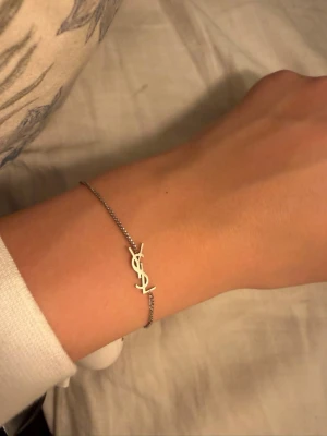 YSL armband - Snyggt och stilrent armband från Yves Saint Laurent i silverfärg. Tunt kedjearmband med den ikoniska YSL-logotypen i mitten. Perfekt för dig som gillar minimalistiska och exklusiva accessoarer.
