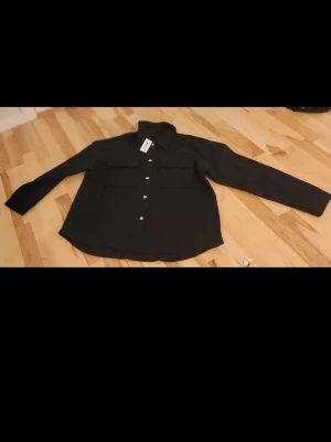 Svart overshirt från New Yorker - Stilren svart overshirt från New Yorker med klassisk krage, knäppning framtill och två stora bröstfickor. Overshirten har raka ärmar och en avslappnad passform. Perfekt att slänga över en t-shirt för en cool look.