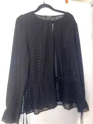 Svart transparent blus med prickar - Snygg svart blus i transparent tyg med diskreta prickar och volang nertill. Långa ärmar med knytband vid ärmslut och rund halsringning. Perfekt att styla med jeans eller kjol för en trendig look.
