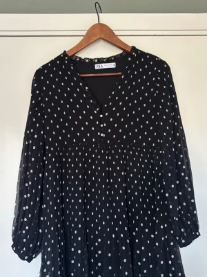 Svart prickig klänning från ZARA - Supersöt svart klänning från ZARA med vita prickar, v-ringning och knappar framtill. Långa, lätt puffiga ärmar och plisserad kjol 
