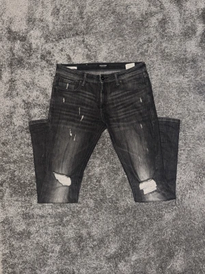 Svarta slitna jeans från Jack & jones - Snygga svarta jeans från Jack & jones med slitningar och hål på knäna. Jeansen har fem fickor, smal passform och cool tvättad look. Perfekta för en trendig streetstyle. Materialet är klassisk denim med stretch för extra komfort.