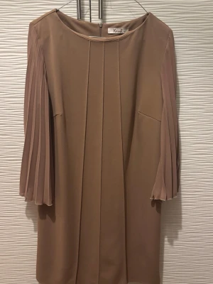 Beige klänning med plisserade ärmar - Stilren beige klänning med raka linjer och plisserade trekvartsärmar. Klänningen har rund halsringning och dragkedja bak. Perfekt för dig som gillar en enkel men elegant look.