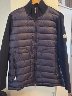 Moncler Cardigan - Snygg svart cardiganjacka från Moncler med quiltad framsida och stickade ärmar samt rygg. Jackan har dragkedja framtill, två fickor med dragkedja och Moncler-logga på ena ärmen. Perfekt för lager-på-lager och en stilren look. 