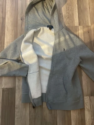 Grå hoodie från Polo Ralph Lauren - Mysig grå hoodie från Polo Ralph Lauren med dragkedja och vit fluffig insida. Klassisk huva, två fickor framtill och broderad logga på bröstet. Perfekt för dig som gillar stilren och bekväm streetwear. Säljer den för att den är för liten på mig nu, den har en avslappnad fit. Nypris ligger på 1000/1200kr. priset kan diskuteras.