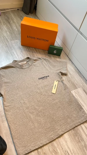 Fear of God Essentials Shirt - Säljer en helt ny Essentials t-shirt från Fear of God i en snygg gråmelerad färg, perfekt nu inför sommaren. Storlek XS men på grund av oversized fit passar den som S. T-shirten har den klassiska loggan på bröstet och den välkända passformen. Alla tags sitter kvar (se bild), så den är aldrig använd. Toppskick!  