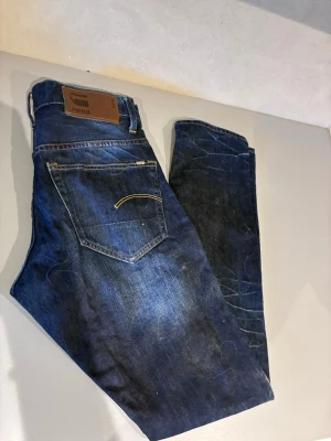 Mörkblå jeans från G-Star RAW - Snygga mörkblå jeans från G-Star RAW med klassisk femficksdesign och kontrastsömmar. Jeansen har en rak passform och detaljerad bakficka med gul söm samt läderpatch med logga i midjan. Perfekta för en trendig och avslappnad look.