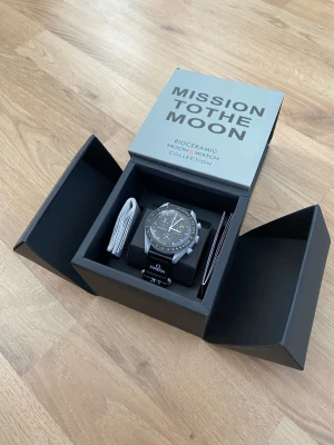 Omega x Swatch MoonSwatch klocka - Säljer en Omega x Swatch MoonSwatch från Bioceramic MoonSwatch Collection. Klockan har svart urtavla med vita detaljer, tachymeter och svart band med Omega-logga. Boetten är i grå biokeramik och designen är inspirerad av rymden.