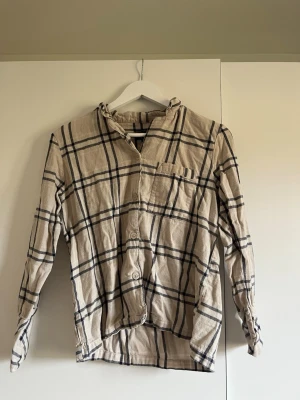 Beige rutig skjorta med knappar - Snygg beige skjorta med svart rutigt mönster och knappar framtill. Skjortan har lång ärm, bröstficka och klassisk krage. Perfekt för en avslappnad och trendig look.