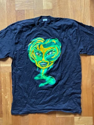 2 vintage Svarta t-shirts street Y2K Japan style  - 2 Unika vintage svarta t-shirts med stort grafiskt tryck av ett färgstarkt ansikte i grönt, gult, rosa och lila på bröstet. T-shirten har rund hals och korta ärmar, perfekt för dig som vill sticka ut med din stil. Storlek L 