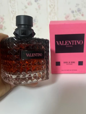 Valentino Donna Born In Roma Intense - Exklusiv parfym från Valentino, Donna Born In Roma Intense. Flaskan är fyrkantig med nitar och har en gradient från transparent till mörkrosa. Materialet är glas och locket är svart med pyramidformade detaljer. Medföljer en rosa kartong med svart text.