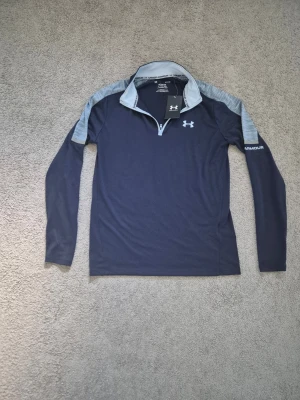 Under Armour - Mörkblå långärmad träningströja från Under Armour med ljusgrå axelpartier och krage. Tröjan har en halv dragkedja framtill och är tillverkad i ett syntetmaterial som andas. Under Armour-logga på bröstet och ärmen.
