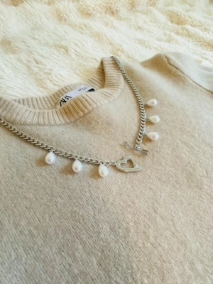 Silverfärgat halsband med pärlor och hjärta - Stilrent halsband silverfärgad rostfritt med droppformade pärlor och ett dekorativt hjärta som låsdetalj. Kedjan är chunky och ger en trendig look, perfekt för att lyfta din outfit.