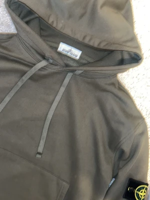 Olivgrön hoodie från Stone Island storlek M - Snygg olivgrön hoodie från Stone Island med klassisk patch på vänster ärm. Hoodien har huva med dragsko, magficka och ribbade muddar. Perfekt för en chill och stilren look. Materialet är mjukt och skönt, troligtvis bomull.
