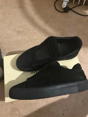 Svarta sneakers från Axel Arigato - Paket ingår. Snygga helsvarta sneakers från Axel Arigato i mocka med minimalistisk design. Skorna har en låg siluett och bred kardborreknäppning istället för klassiska snören. Perfekta för dig som gillar stilrena och trendiga skor. 