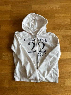 Vit hoodie från Hollister med tryck - Vit hoodie från Hollister med dragkedja och svart tryck framtill med texten 'Hollister 22 California'. Mjuk bomullskänsla, huva med dragsko och två fickor framtill. Perfekt för en avslappnad och stilren look. Priset går att diskutera och jag skickar snabbt. Storleken är M men sitter mer som S