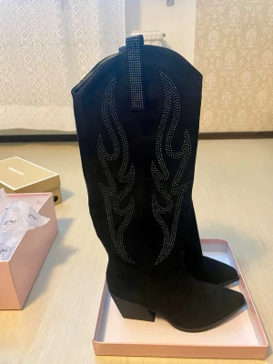 *NY*Swedish Santiags with black rhinestones stl 37 - Nya i kartong fr Stephan, Paris.  Western style svarta mockaboots i stl 37 (23,7 cm innermått). Sitter enligt mig snyggast på ett lite smalare ben/vad. Rhinestones på utsidan och helsvart på insidan, ofodrade. Klack 7 cm. Nypris: 599 sek Pris nu: 399 sek. Our suede western-style cowboy boots are designed to add an authentic touch to your footwear collection. Whether you pair them with casual jeans or a bohemian dress, these boots offer a unique and fashionable combination. 