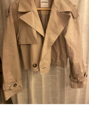 Kort beige trenchcoat från Bershka - Stilren kort trenchcoat i beige från Bershka med breda slag, axelklaffar och knappar vid ärmslut. Jackan har en klassisk design med modern twist och är tillverkad i ett lätt tyg som passar perfekt till lager-på-lager-stil.