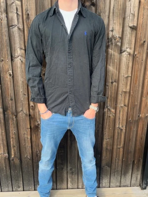 Svart skjorta från Ralph Lauren - Klassisk svart skjorta från Ralph Lauren med blå broderad logga på bröstet. Skjortan har lång ärm, knappar framtill och är tillverkad i bomull. Perfekt för en stilren look. Stolrek M och mycket bra skick! Vi kan stryka skjortan innan leverans om så önskas😊 skick 10/10, hör av er vid frågor // studio II Resell 💙
