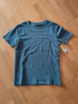 Blå t-shirt med bröstficka  - Säljer en blå  i storlek S. T-shirten har en enkel design med rund halsringning och en bröstficka på vänster sida. Materialet känns mjukt och lite strukturerat, perfekt för en avslappnad stil.